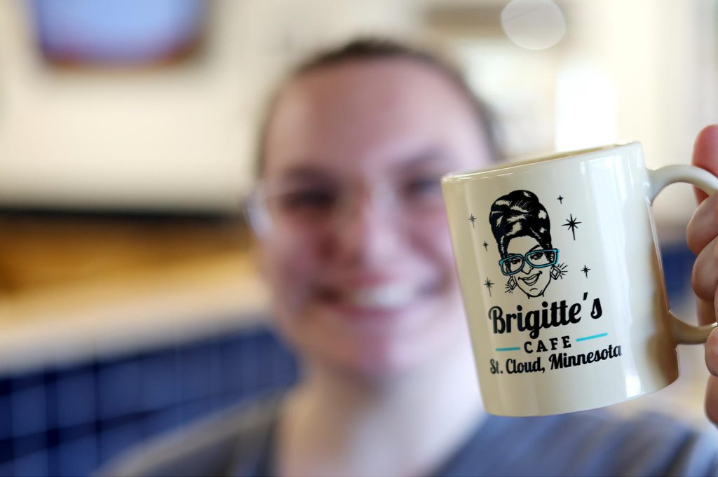 Blurred woman in background holding up a coffee mug in focus with the Brigitte's Cafe logo on it.